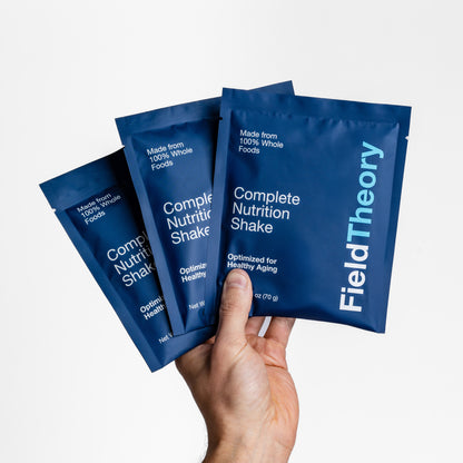 Hand holding three blue Field Theory Complete Nutrition Shake single-serving Travel Packs packages against a white background